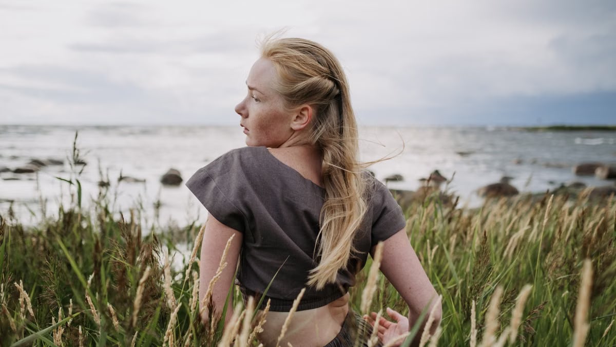 A woman with long blonde hair sitting peacefully in grass by the ocean, showcasing a summer vibe.